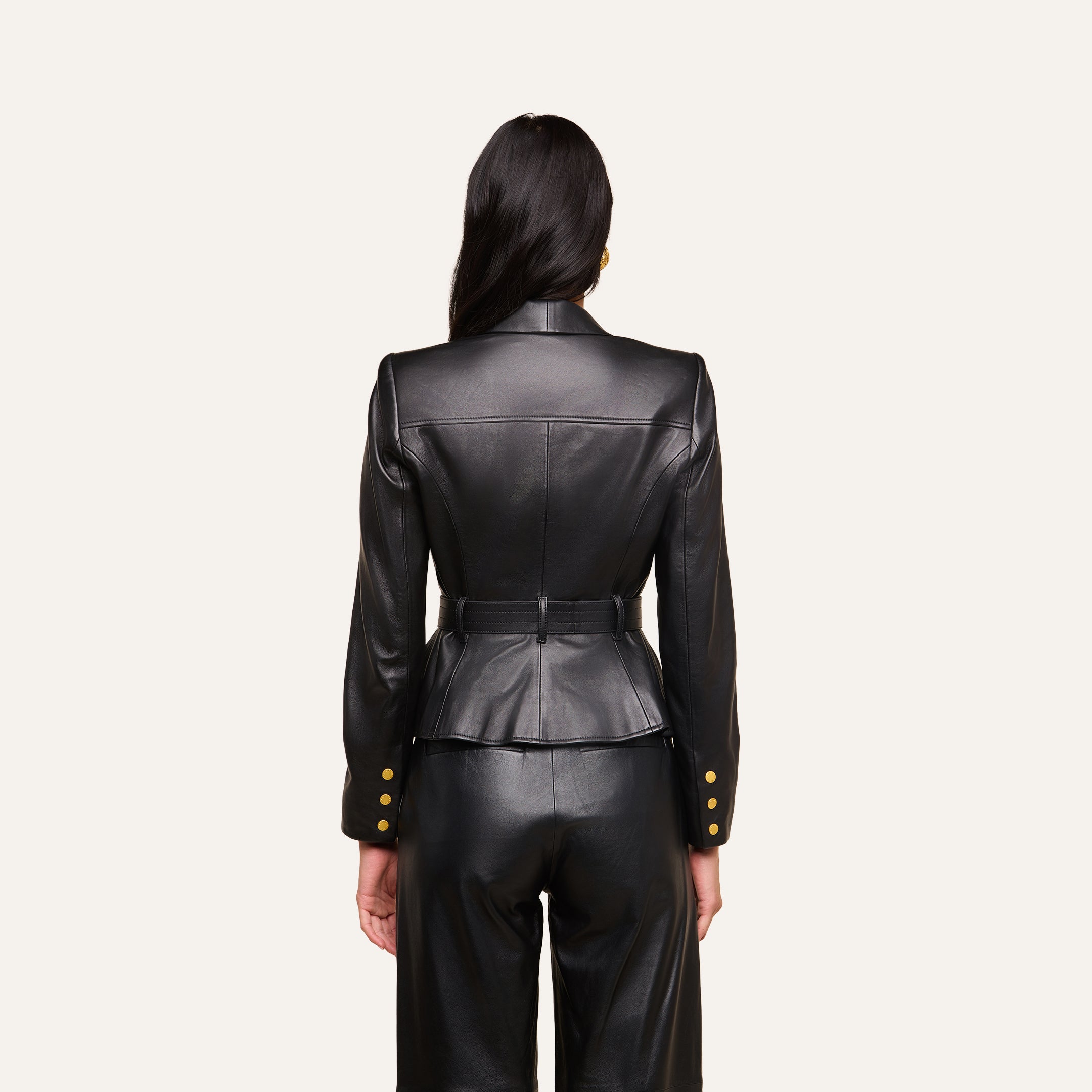 A woman with long dark hair, seen from behind, wears the fitted Wisteria Leather Blazer in black, paired with matching pants. The blazer features gold buttons on the cuffs and a belted waist. The background is plain and light-colored.