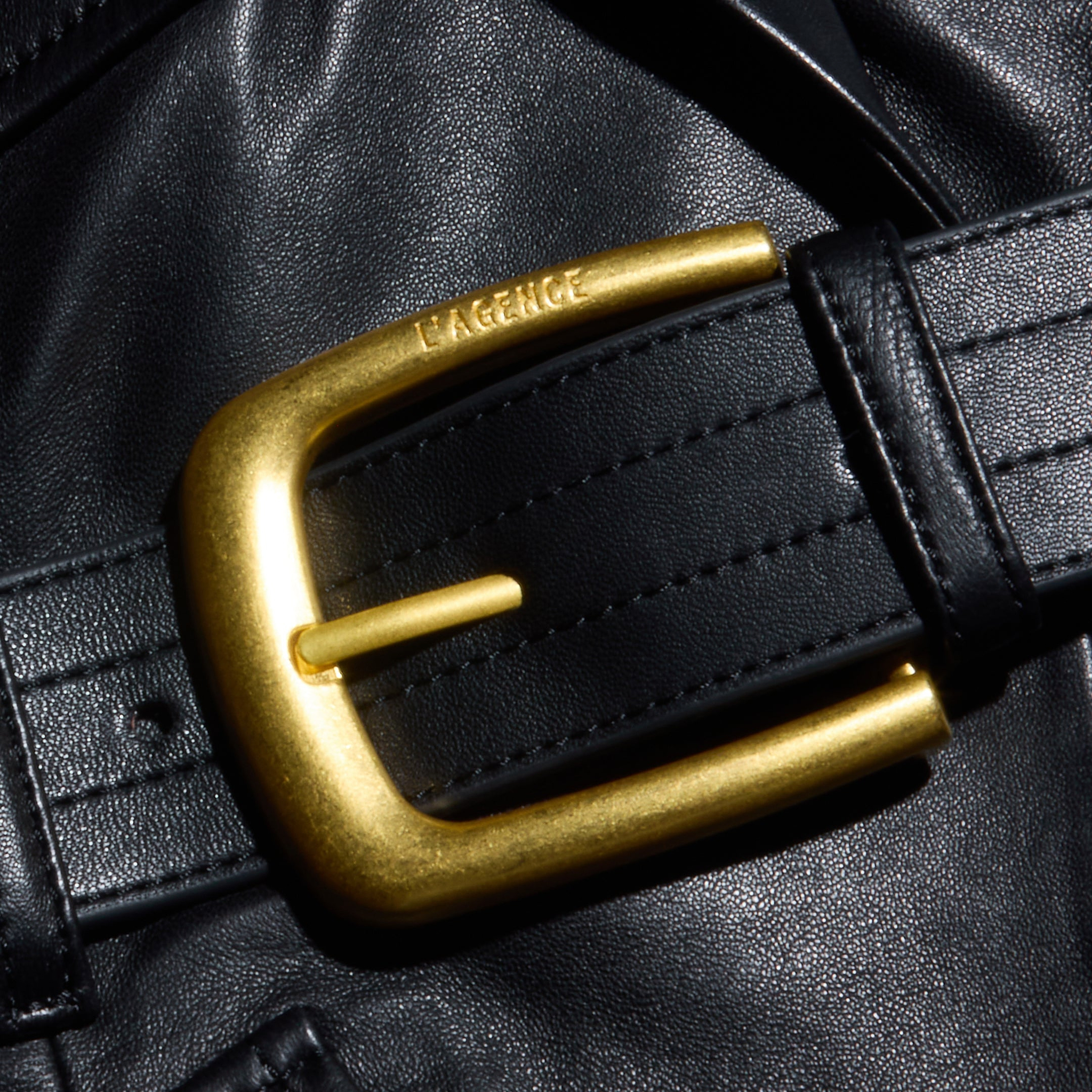 A close-up of a shiny gold belt buckle engraved with the word LAGENCE adorns a black leather belt set against the luxurious black leather background of the Wisteria Leather Blazer, evoking sophisticated, tailored style.