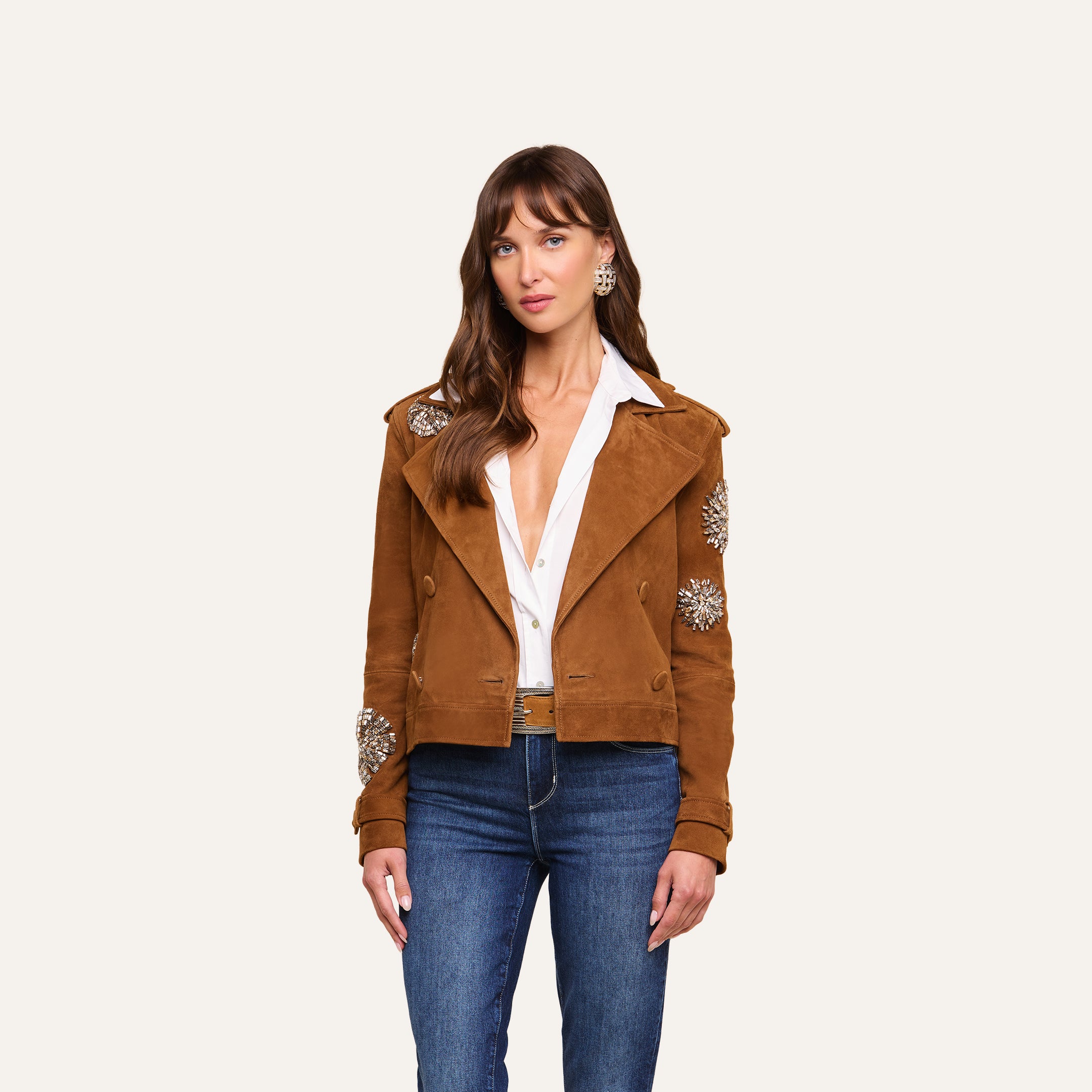 A woman stands against a plain light background, wearing the Xoey Cropped Crystal Trench Coat over a white open-collar shirt, styled with blue jeans and statement earrings, showcasing a look inspired by Fall 2026 Fashion Week.