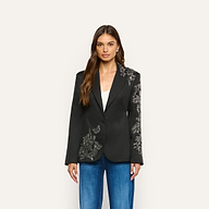 A woman wearing the Clementine Embellished Blazer, a black twill jacket with silver beaded floral embroidery on one sleeve and the opposite hip, paired with blue jeans and a white top.