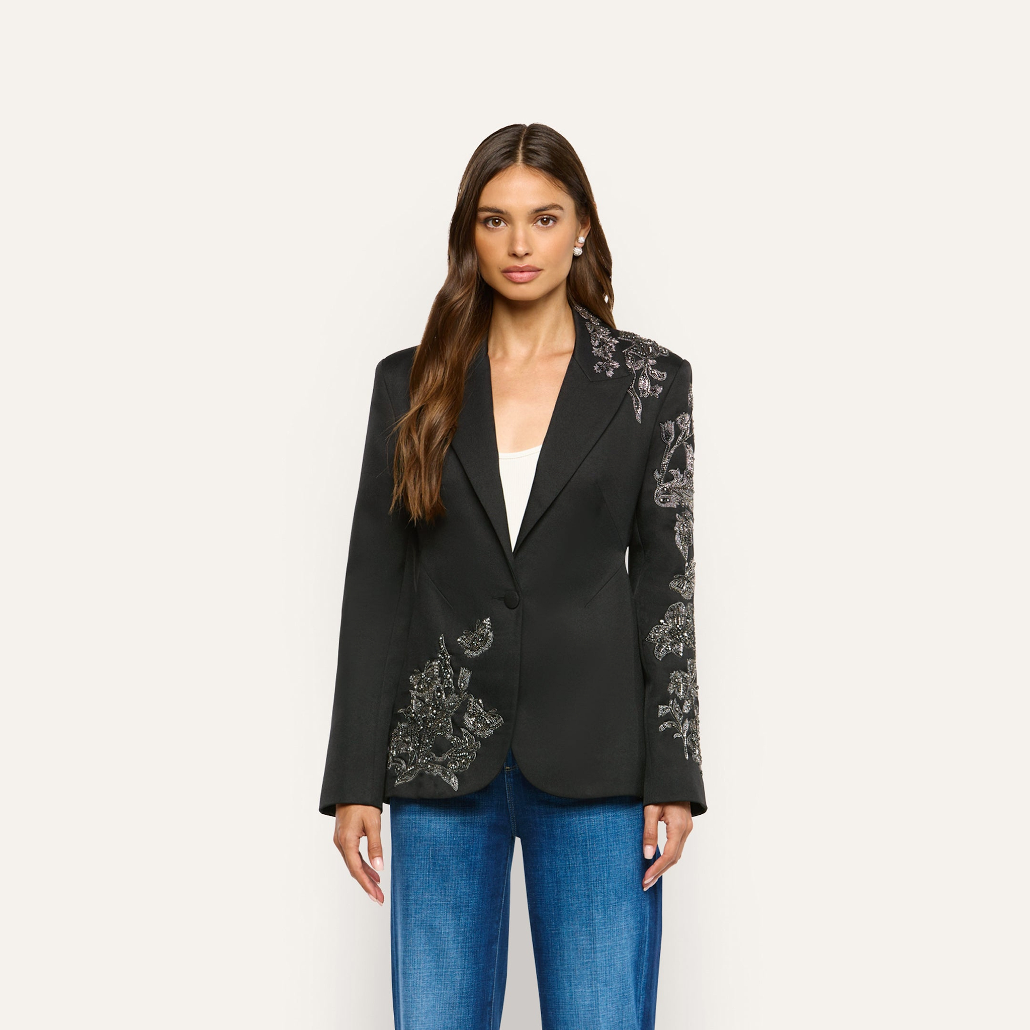 A woman wearing the Clementine Embellished Blazer, a black twill jacket with silver beaded floral embroidery on one sleeve and the opposite hip, paired with blue jeans and a white top.