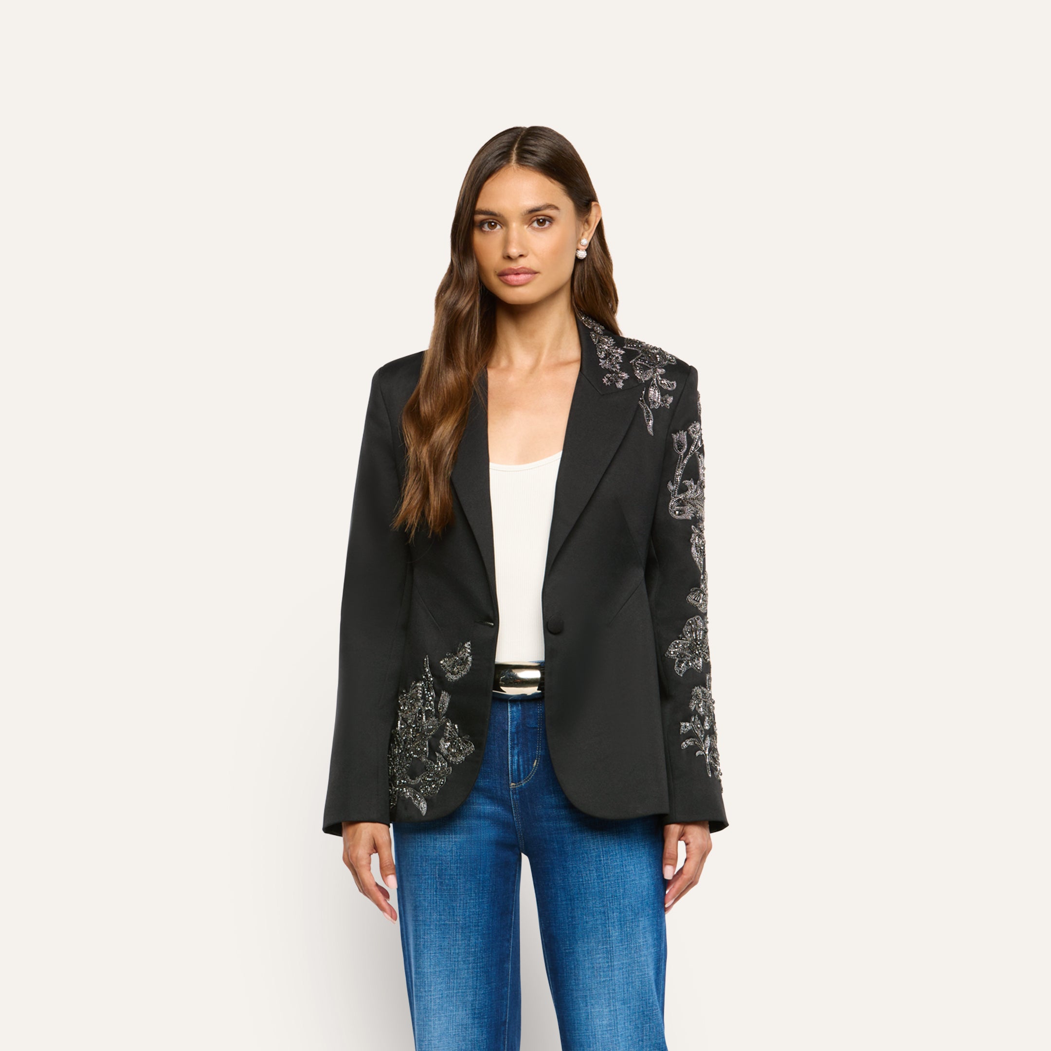 A woman wears a black, crystal-embellished Clementine Embellished Blazer with silver floral embroidery on one shoulder, sleeve, and the opposite hip, paired with a white tank top and blue jeans.