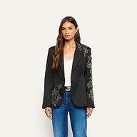 A woman wears a black, crystal-embellished Clementine Embellished Blazer with silver floral embroidery on one shoulder, sleeve, and the opposite hip, paired with a white tank top and blue jeans.