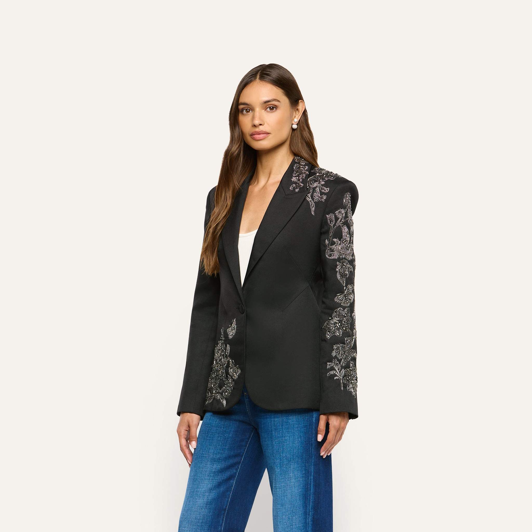 A woman wears a black twill Clementine Embellished Blazer featuring intricate floral embroidery on the shoulders, sleeves, and front pocket area, paired with blue jeans.