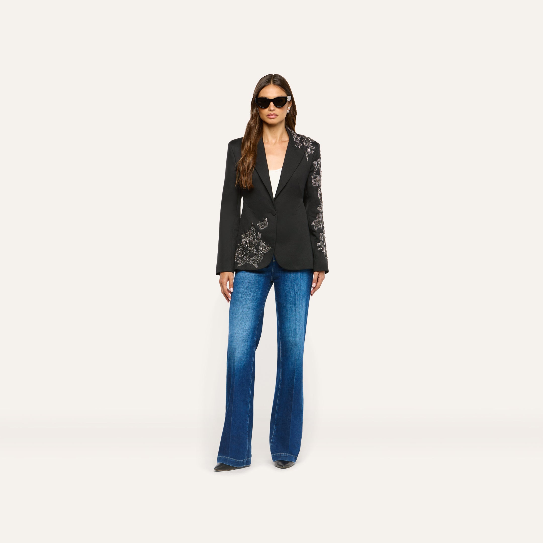 A woman wearing a black Clementine Embellished Blazer with crystal-embellished floral embroidery on the shoulder and hip, paired with blue flared jeans and sunglasses.