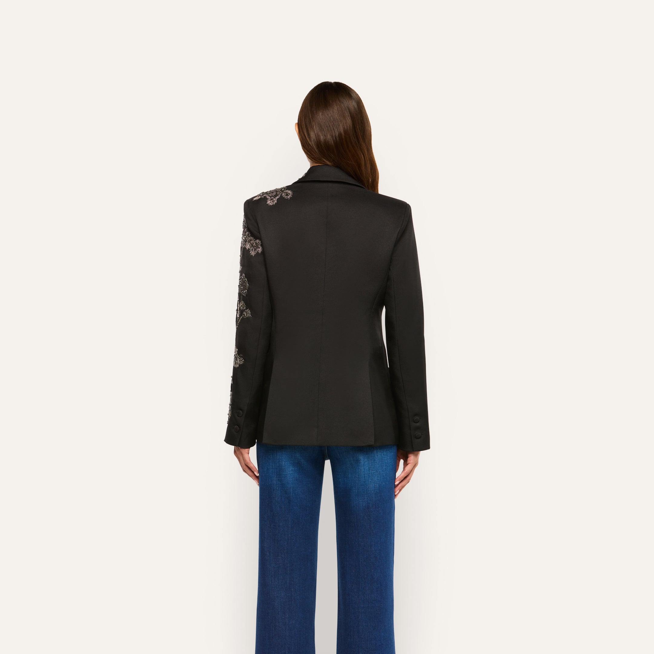 Back view of the black twill Clementine Embellished Blazer with floral embroidery on the right shoulder and sleeve, worn with blue jeans.