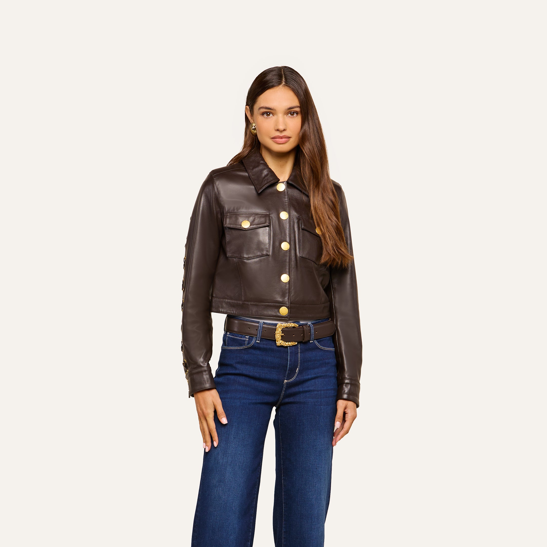 A woman stands facing forward, wearing the Luci Fringed Leather Jacket—a dark brown biker style featuring gold buttons and front pockets—paired with blue jeans and a black belt with a gold buckle. She has long brown hair and a neutral expression.