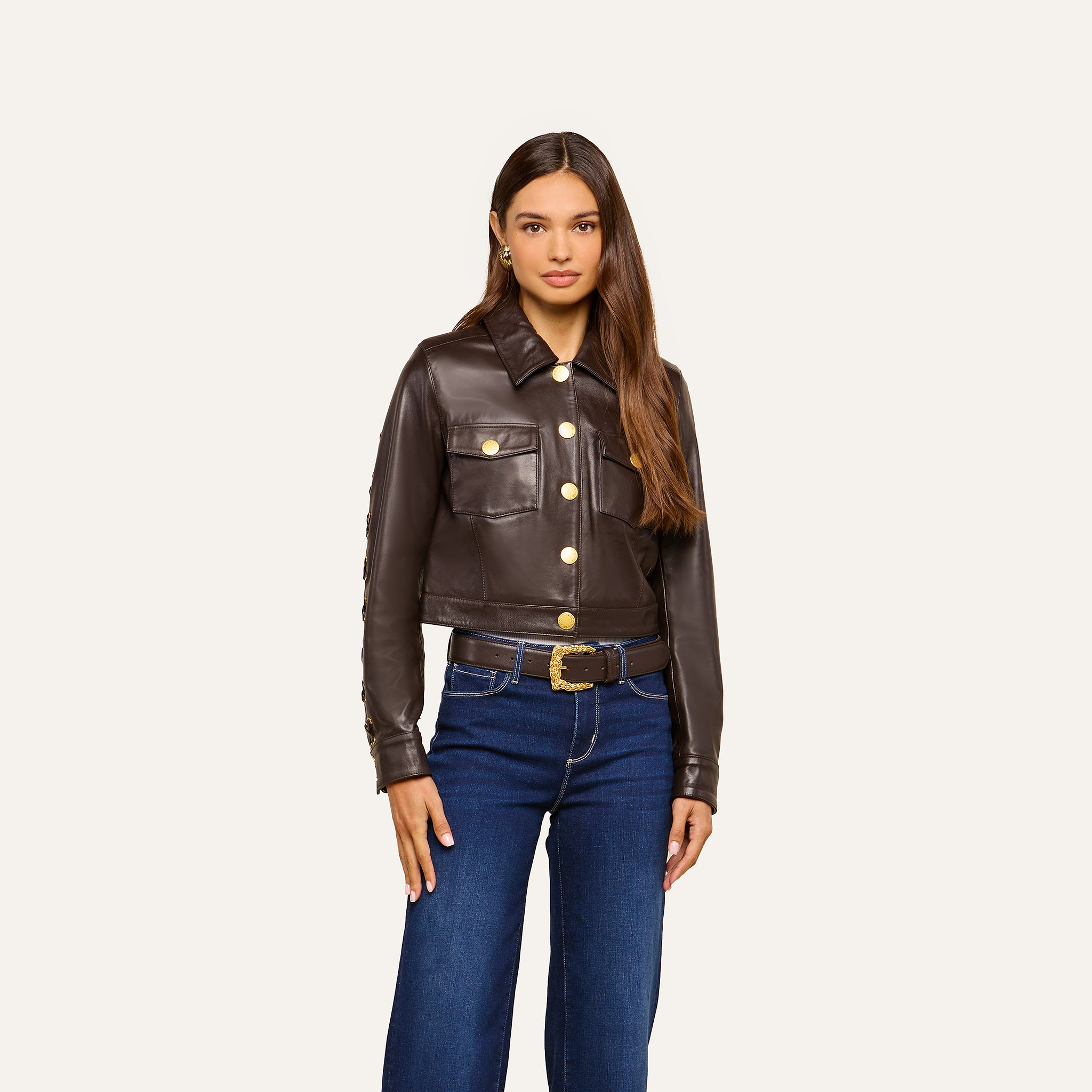 A woman stands facing forward, wearing the Luci Fringed Leather Jacket—a dark brown biker style featuring gold buttons and front pockets—paired with blue jeans and a black belt with a gold buckle. She has long brown hair and a neutral expression.