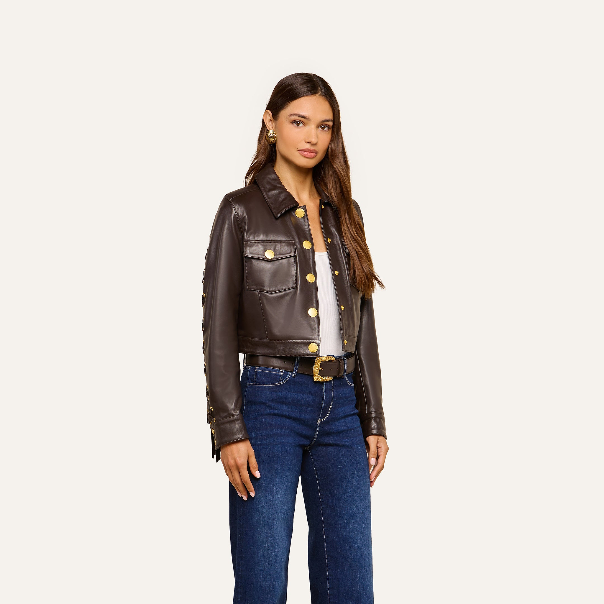 A woman stands facing forward, wearing a Luci Fringed Leather Jacket in dark brown leather with gold buttons, a white top, blue jeans, a black belt with a gold buckle, and gold earrings against a plain, light background.