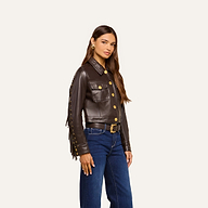 A woman stands confidently against a plain light background, wearing the Luci Fringed Leather Jacket with gold buttons, paired with blue jeans and gold earrings. Her long brown hair cascades over her shoulders as she looks directly at the camera.