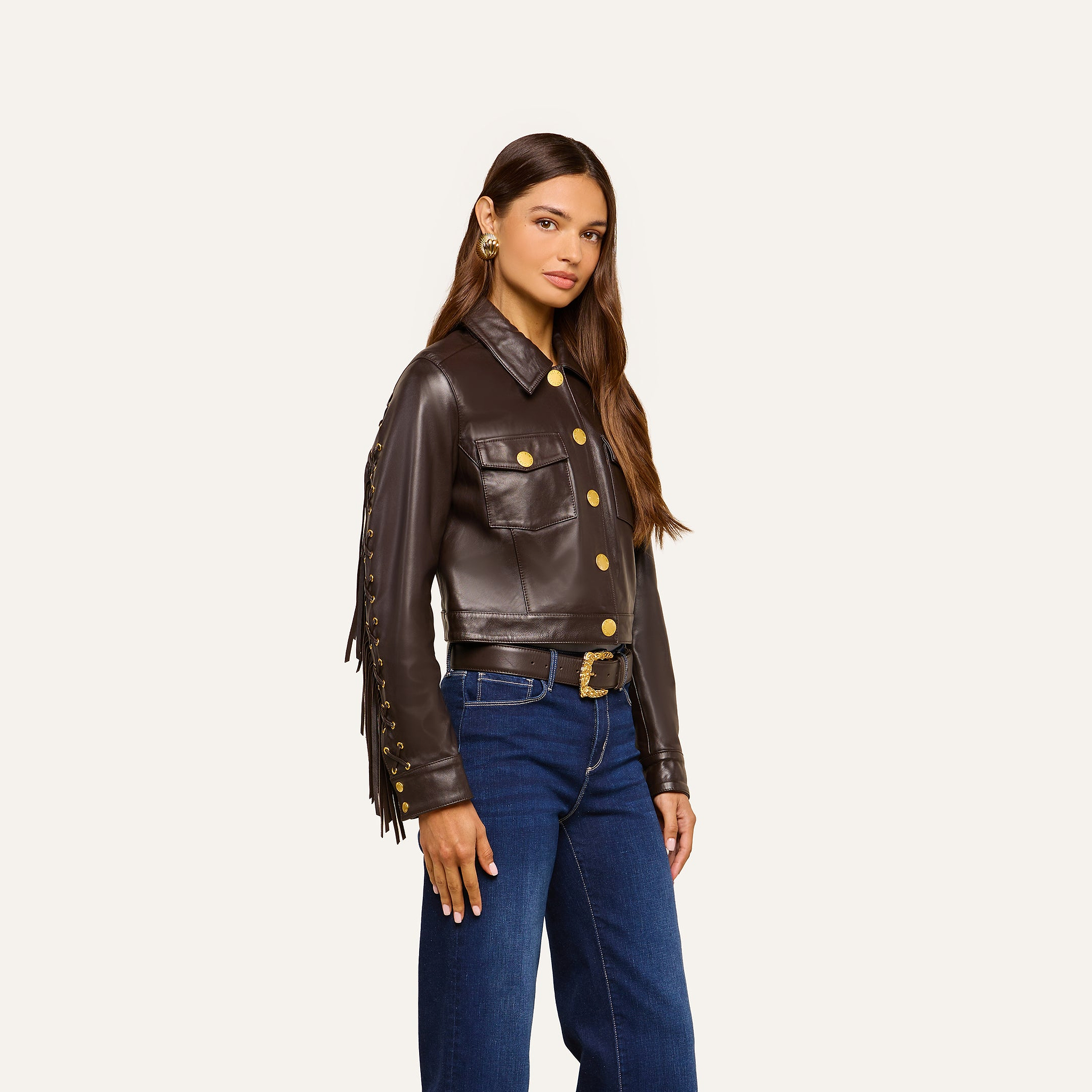 A woman stands confidently against a plain light background, wearing the Luci Fringed Leather Jacket with gold buttons, paired with blue jeans and gold earrings. Her long brown hair cascades over her shoulders as she looks directly at the camera.