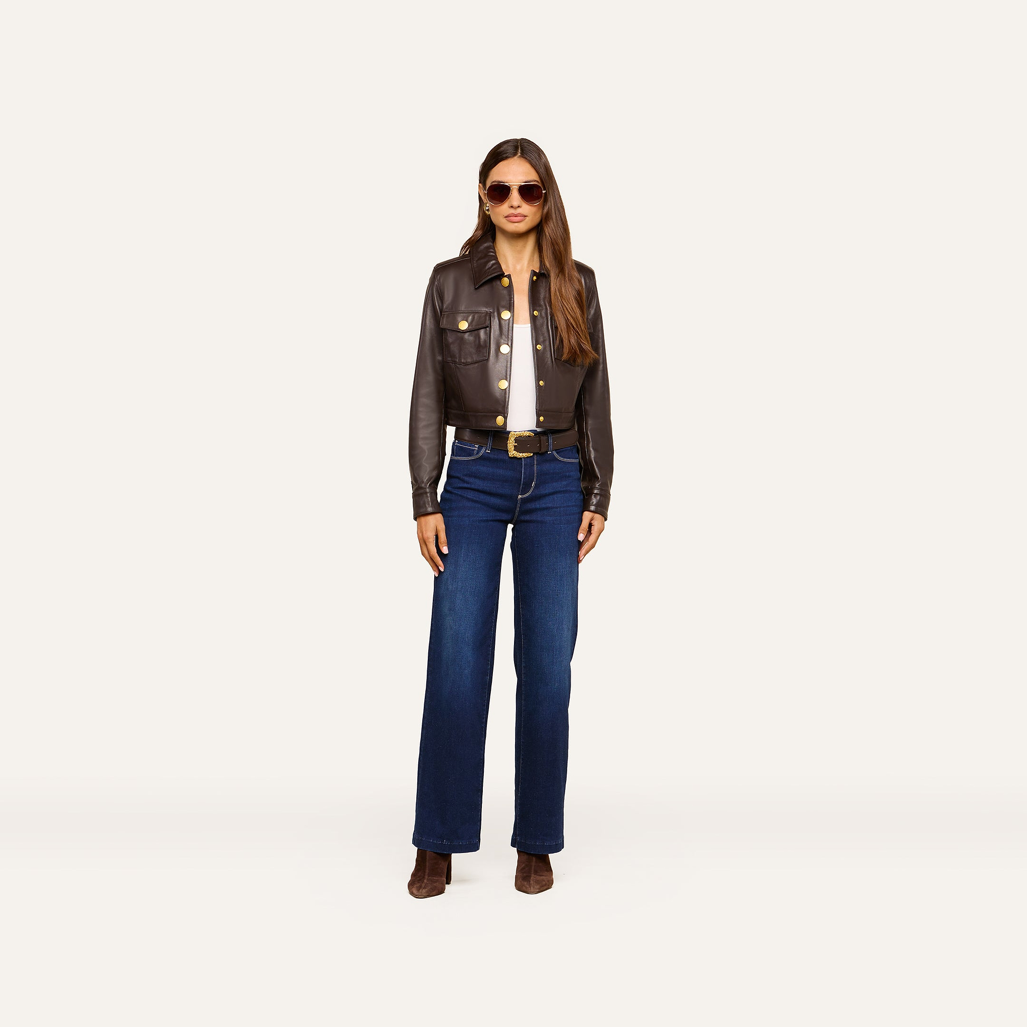 Wearing the Luci Fringed Leather Jacket, a woman stands against a plain white background, styled with sunglasses, a white top, blue high-waisted jeans, a belt, and brown boots.