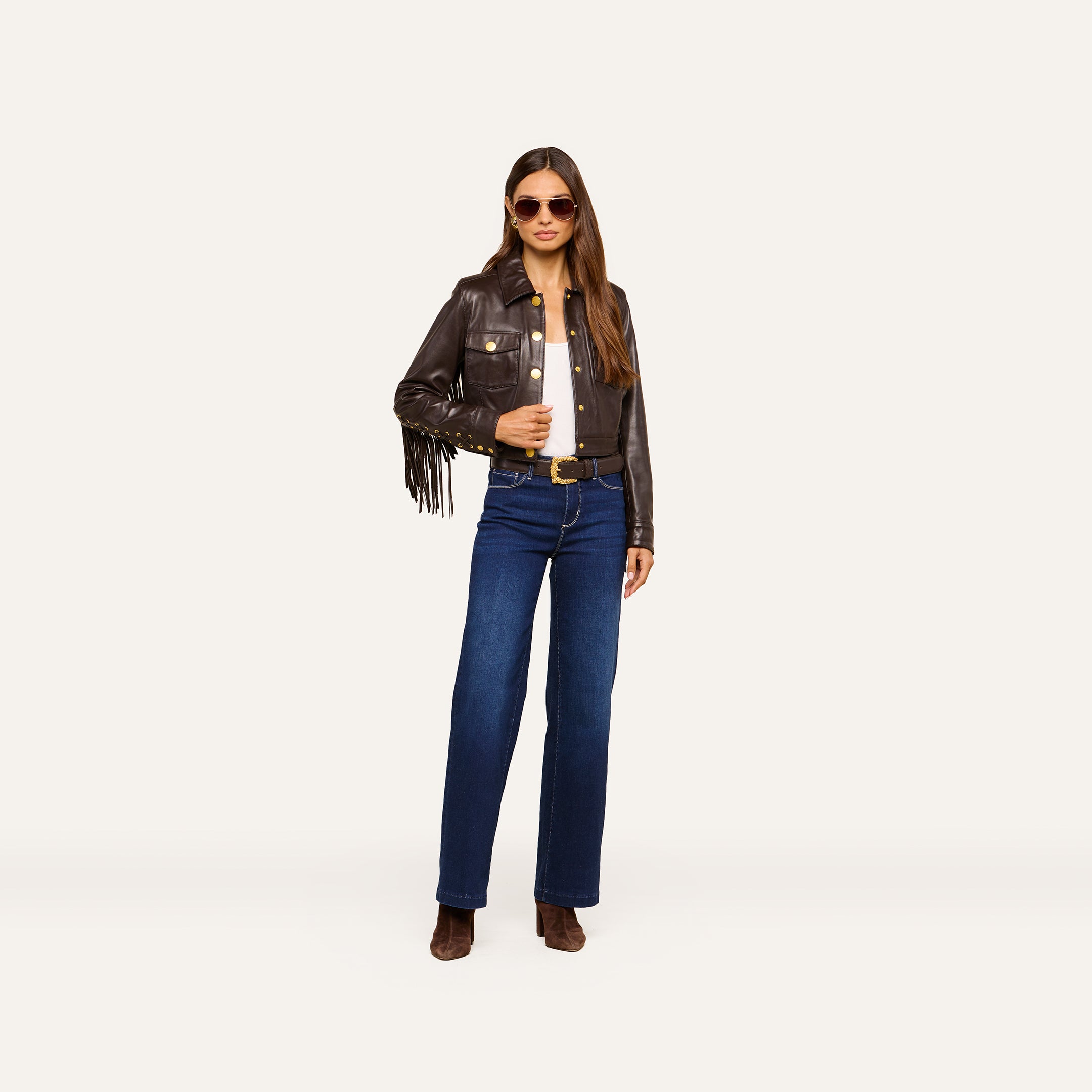 Wearing the Luci Fringed Leather Jacket, a woman in sunglasses, blue jeans, and brown boots stands confidently against a plain light background.