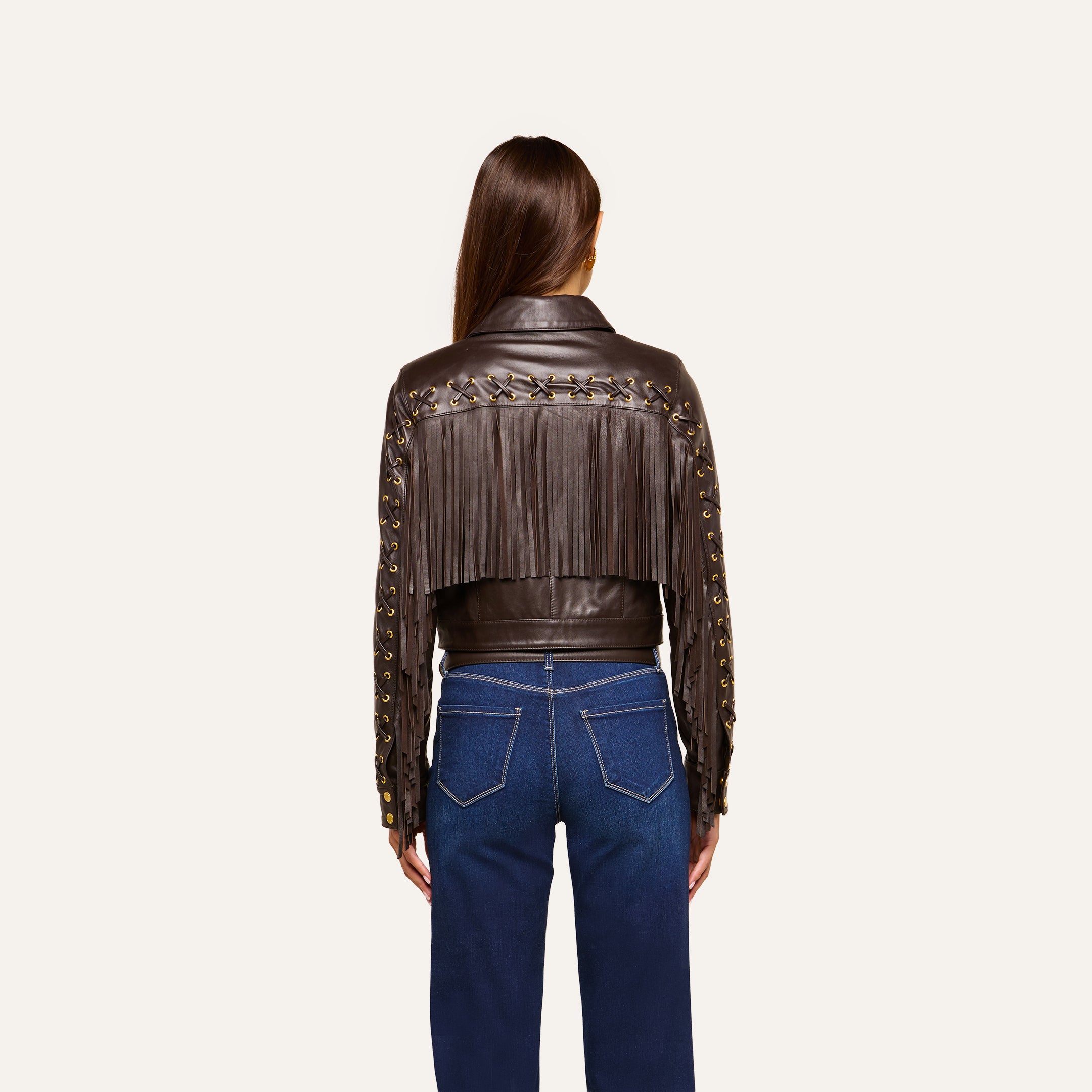 A person with long brown hair, seen from behind, wears the Luci Fringed Leather Jacket with grommet details, paired with blue jeans and standing against a plain light background.