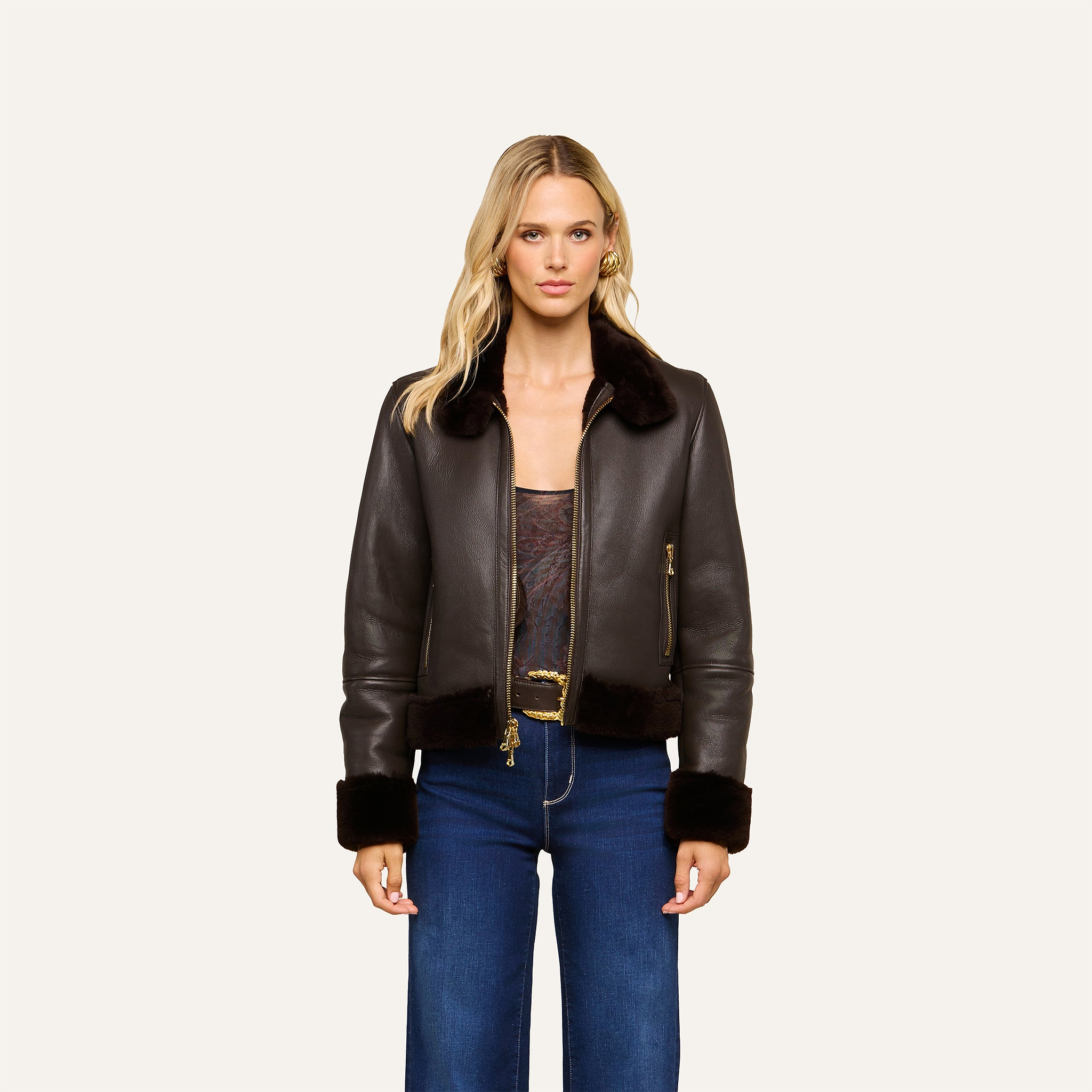 A woman stands against a plain background wearing the Glacia Shearling Jacket—a cropped leather piece with a fur collar and cuffs—paired with a sheer top and blue jeans.