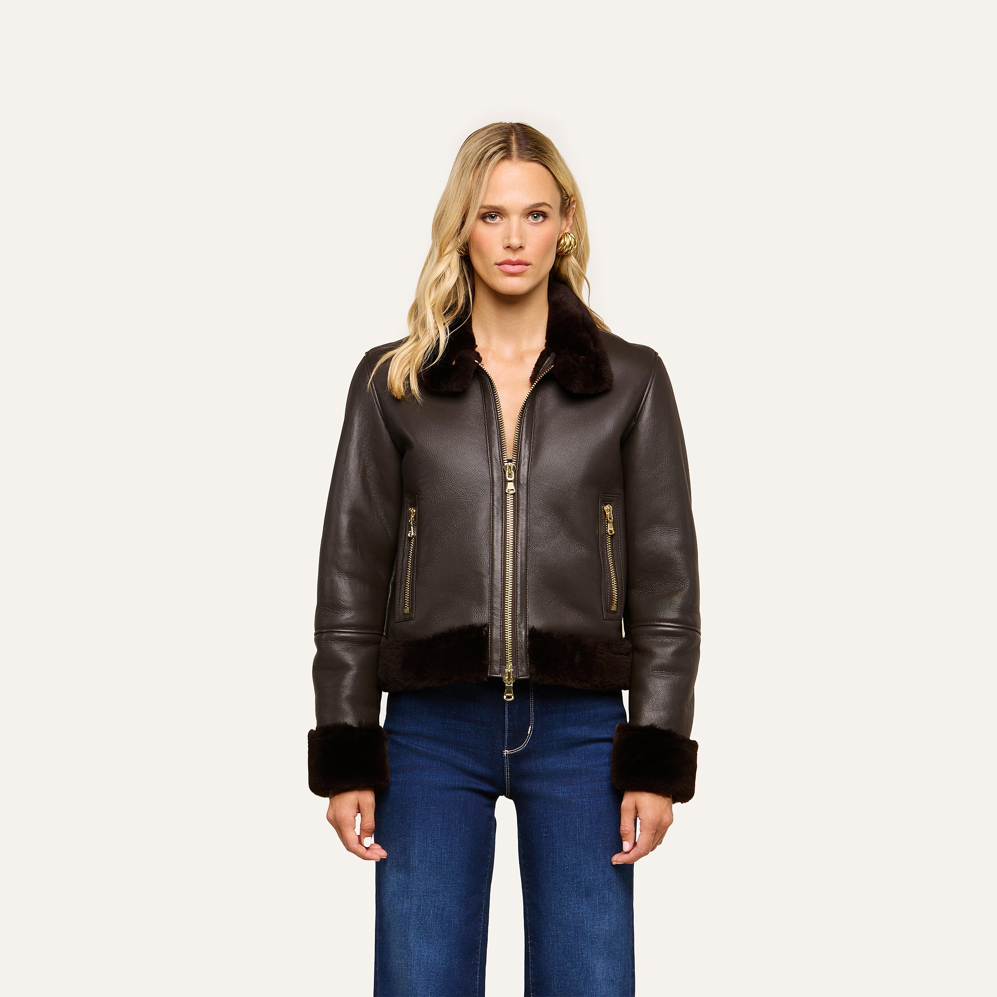 A person wearing the Glacia Shearling Jacket—a dark brown aviator style with fur trim—paired with blue jeans stands facing forward against a plain, light background.