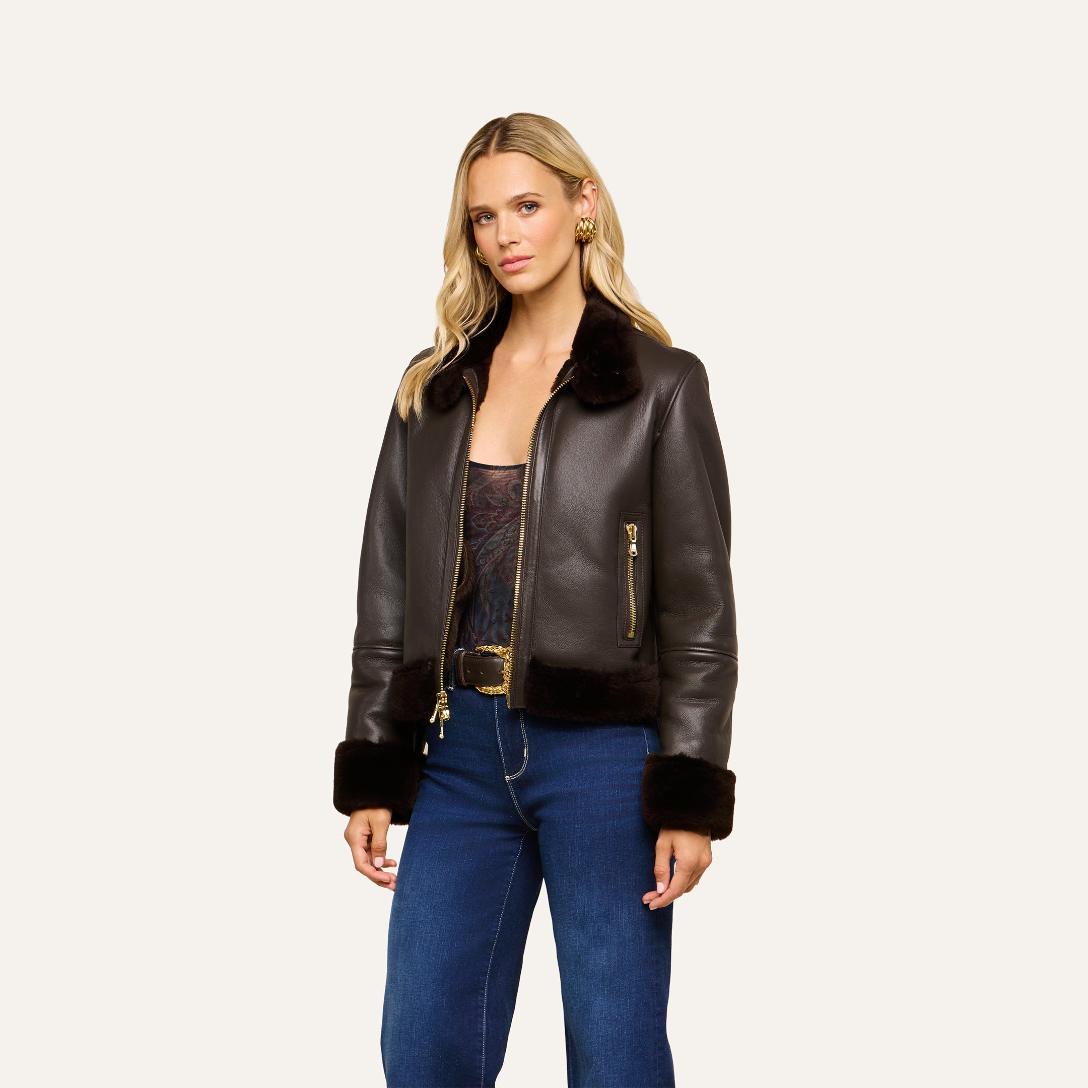 A woman with blonde hair wears the Glacia Shearling Jacket, a dark brown aviator style, over a patterned top and blue jeans, standing against a plain light background.