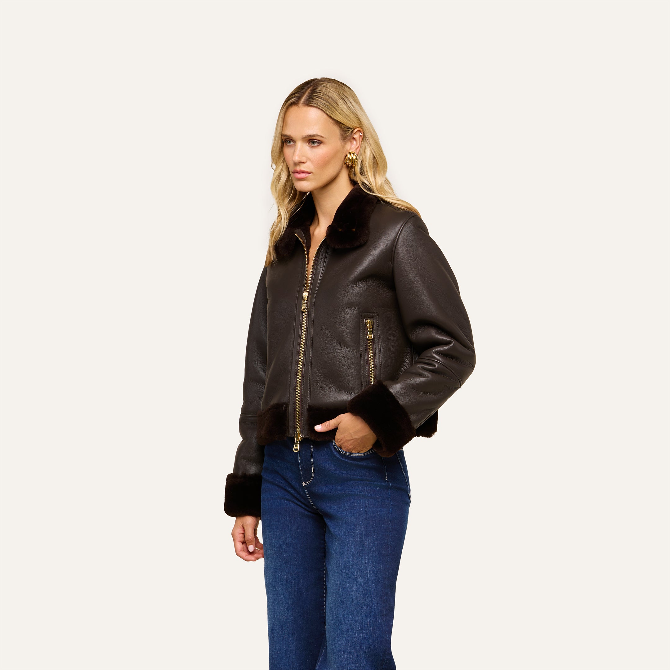 A woman with long blonde hair wears the Glacia Shearling Jacket—a dark brown aviator style with a zip front—paired with blue jeans, standing against a plain light background.