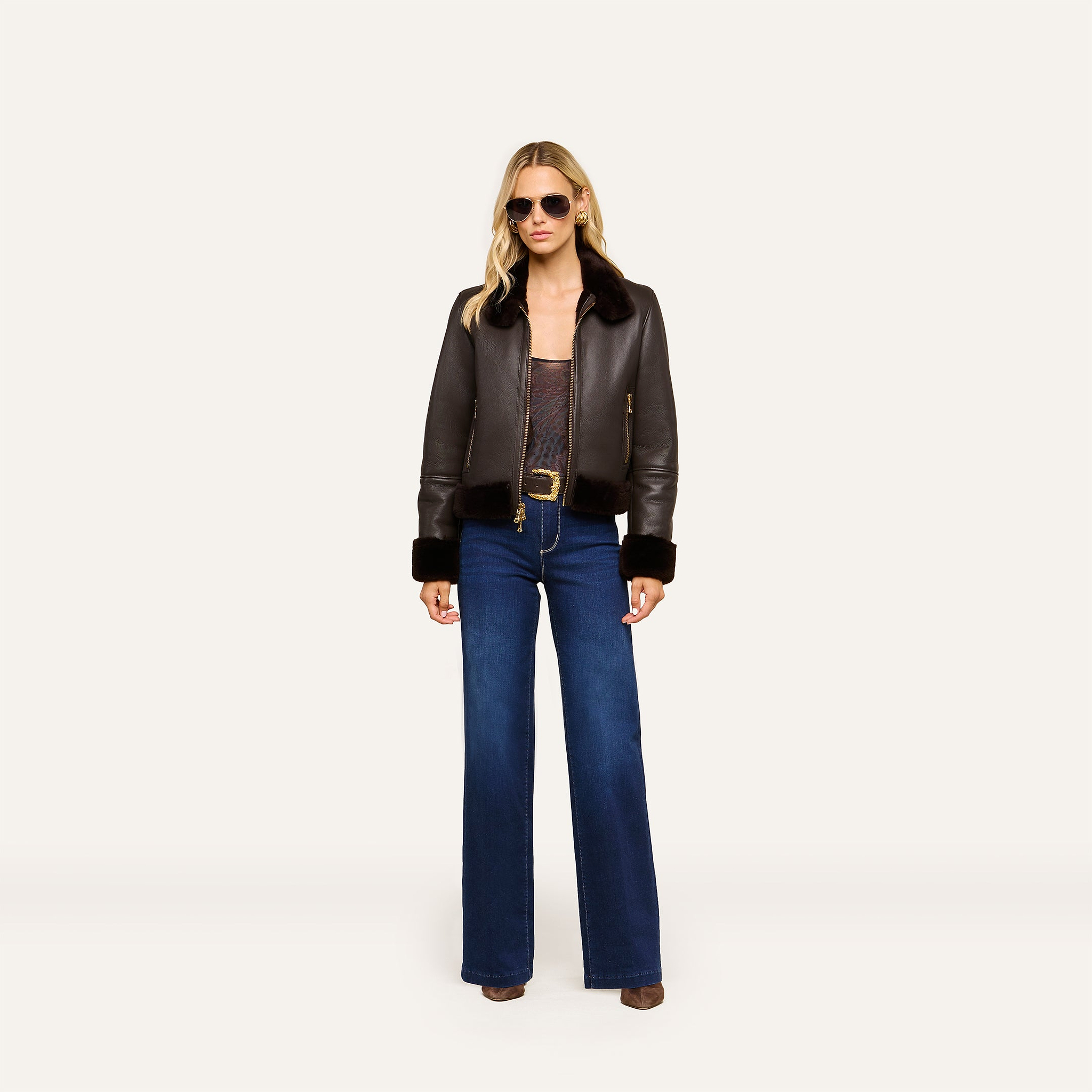 A woman wearing sunglasses, the Glacia Shearling Jacket, a sheer top, blue flare jeans, and brown shoes stands confidently against a plain white background.