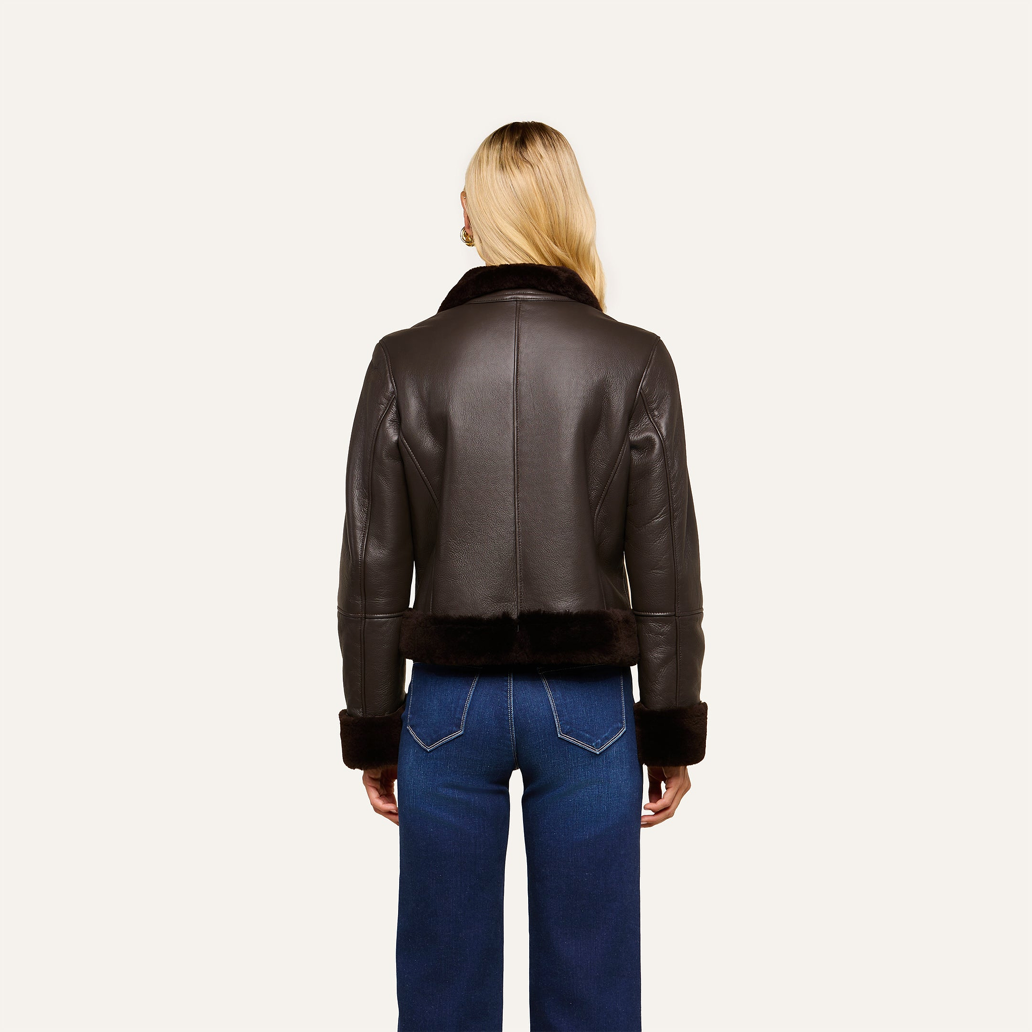 A person with long blonde hair, seen from behind, wears the Glacia Shearling Jacket in dark brown with fur trim and blue jeans. The background is plain and light.
