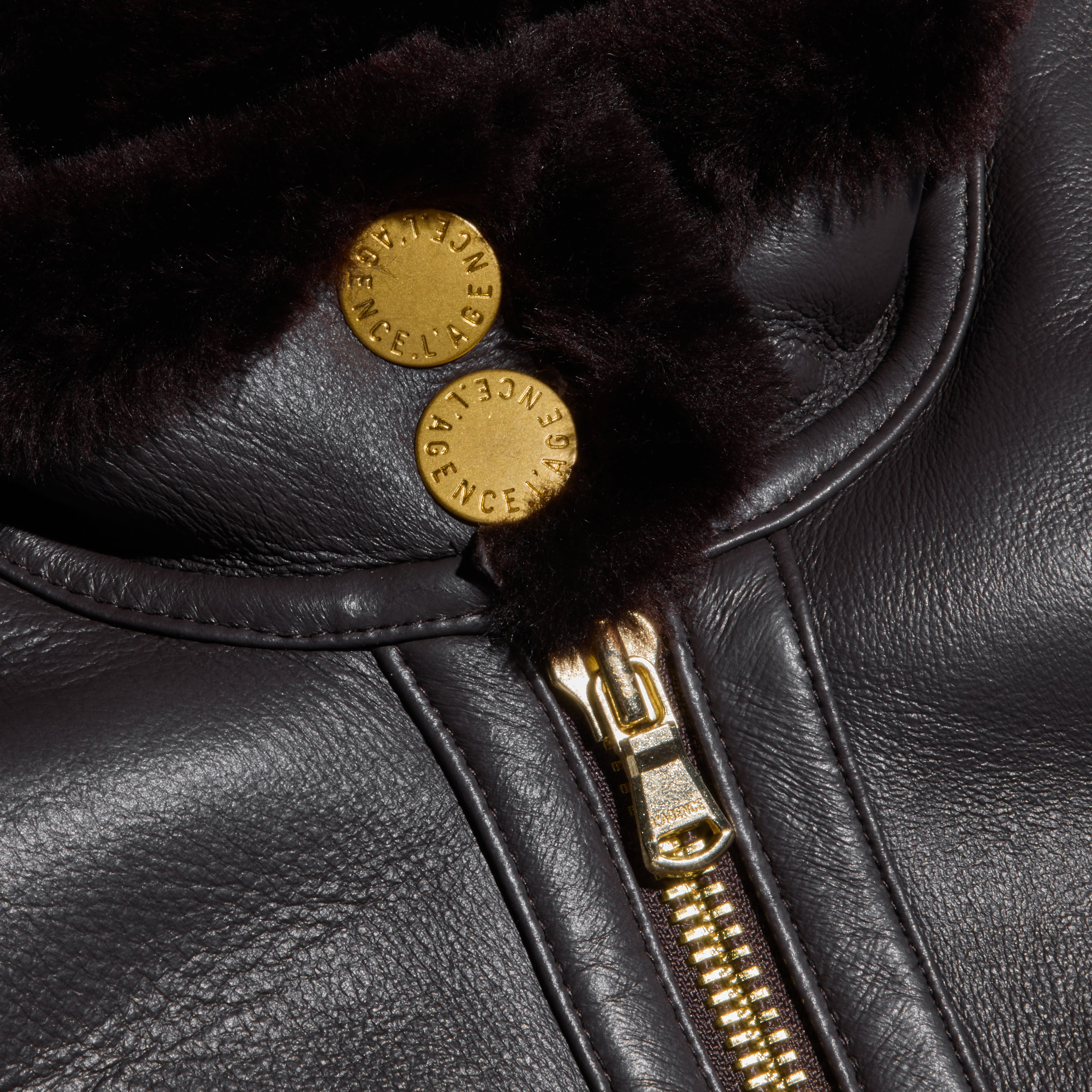 Close-up of the Glacia Shearling Jacket featuring a cropped leather design, gold zipper, brown fur lining, and two brass buttons engraved with text.