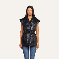 A woman stands facing forward, wearing the Ignis Puffer Vest—a sleeveless, belted vegan leather piece perfect for transitional outerwear—layered over a black top and blue jeans against a plain light background.