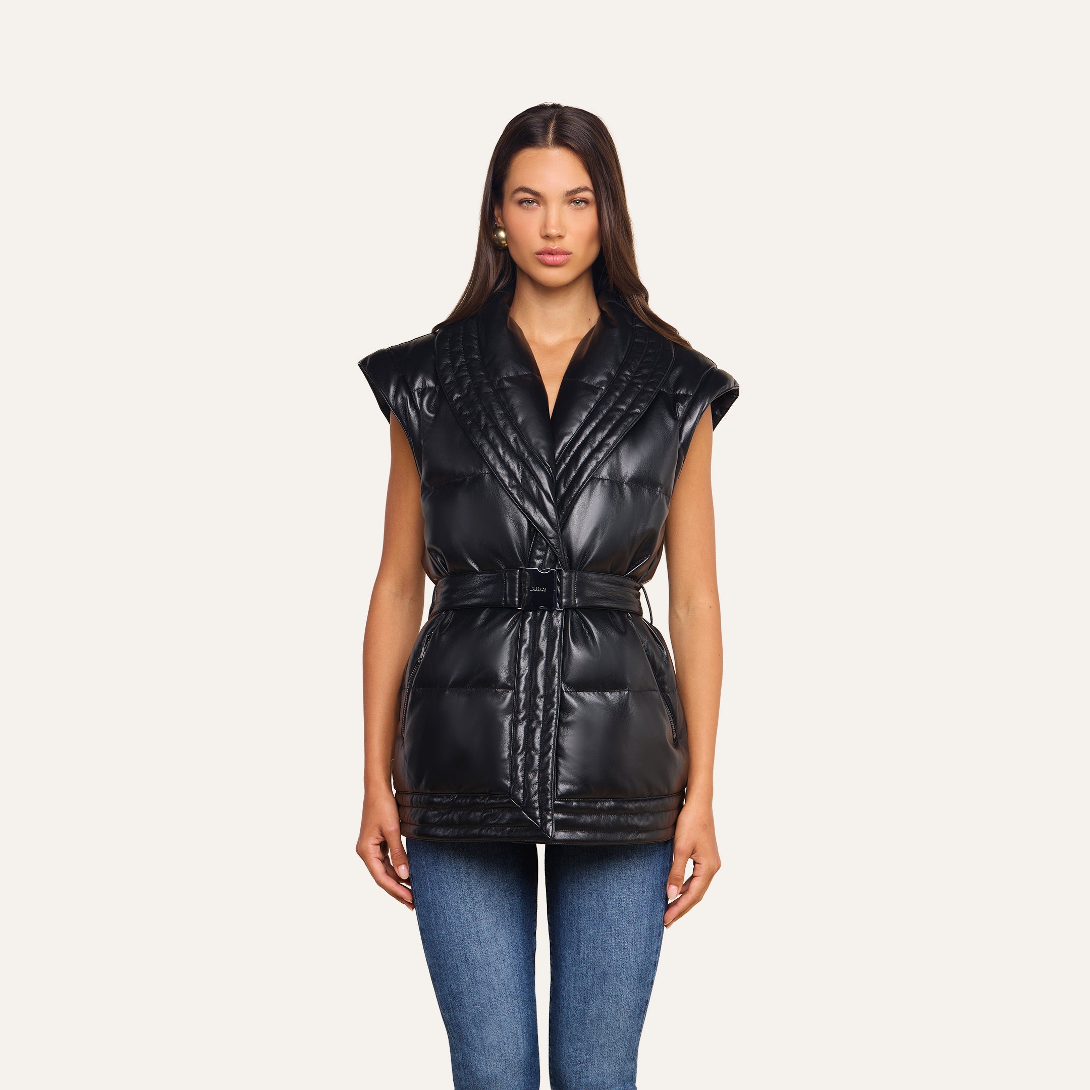 A woman stands facing forward, wearing the Ignis Puffer Vest—a sleeveless, belted vegan leather piece perfect for transitional outerwear—layered over a black top and blue jeans against a plain light background.