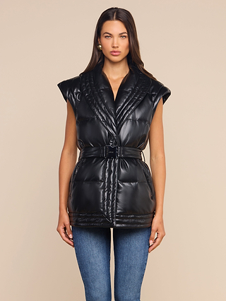 A woman wearing the Ignis Puffer Vest—a black, belted, quilted style featuring cap sleeves and a subtle shawl collar—pairs it with blue jeans as she stands facing forward against a plain beige background, her expression neutral. This transitional outerwear piece adds an elegant touch to her look.