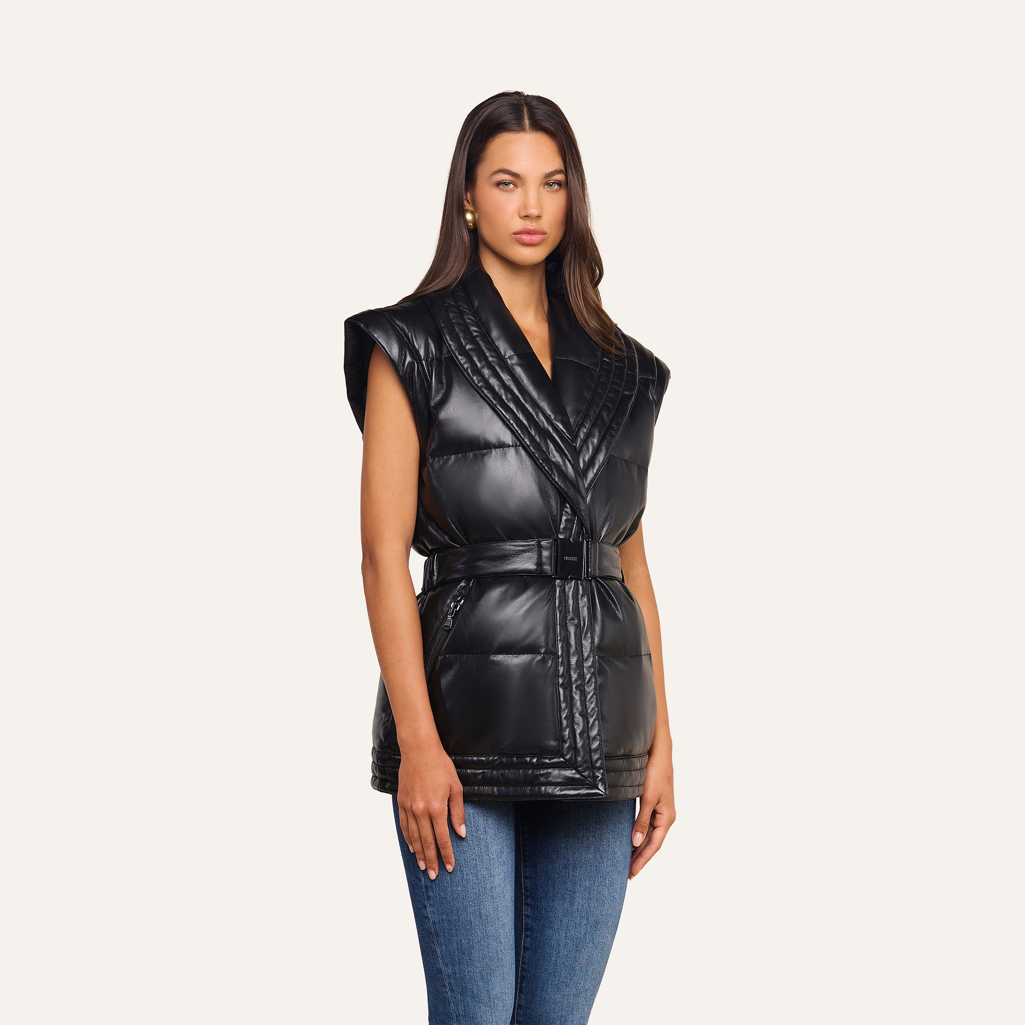 A woman stands facing forward against a plain light background, wearing the Ignis Puffer Vest— a black, sleeveless, quilted vegan leather vest with a belt— styled over a black top and blue jeans. This stylish piece serves as perfect transitional outerwear.