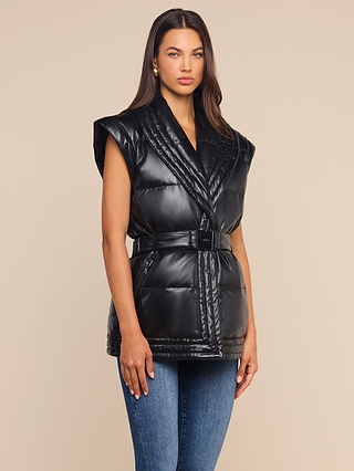 A woman stands in front of a beige background, wearing the Ignis Puffer Vest—a black, belted, sleeveless vegan leather piece—layered over a black top and blue jeans. With her long dark hair and gold earrings, this look is perfect for transitional outerwear.