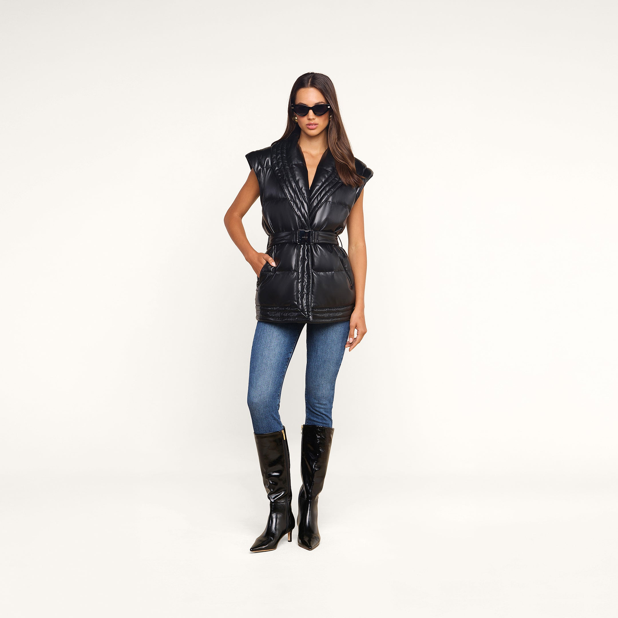 A woman in sunglasses wears the Ignis Puffer Vest—a black vegan leather belted puffer vest with a shawl collar—styled with blue jeans and black knee-high boots, standing against a plain white background.
