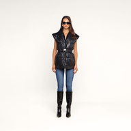 Wearing sunglasses, blue skinny jeans, and black knee-high boots, a woman stands facing forward against a plain white background in the Ignis Puffer Vest—a black vegan leather belted puffer with a shawl collar. This look is perfect for transitional outerwear.