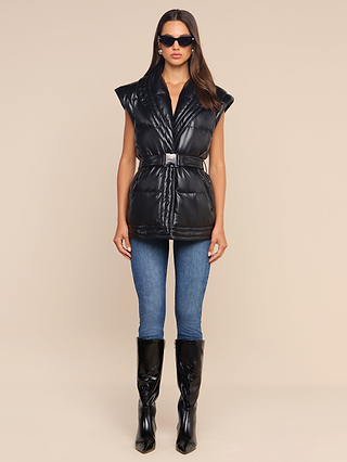 A woman stands facing forward against a plain beige background, wearing the Ignis Puffer Vest—a belted black vegan leather piece with cap sleeves—paired with blue jeans, black knee-high boots, and sunglasses. This sophisticated vest serves as ideal transitional outerwear.