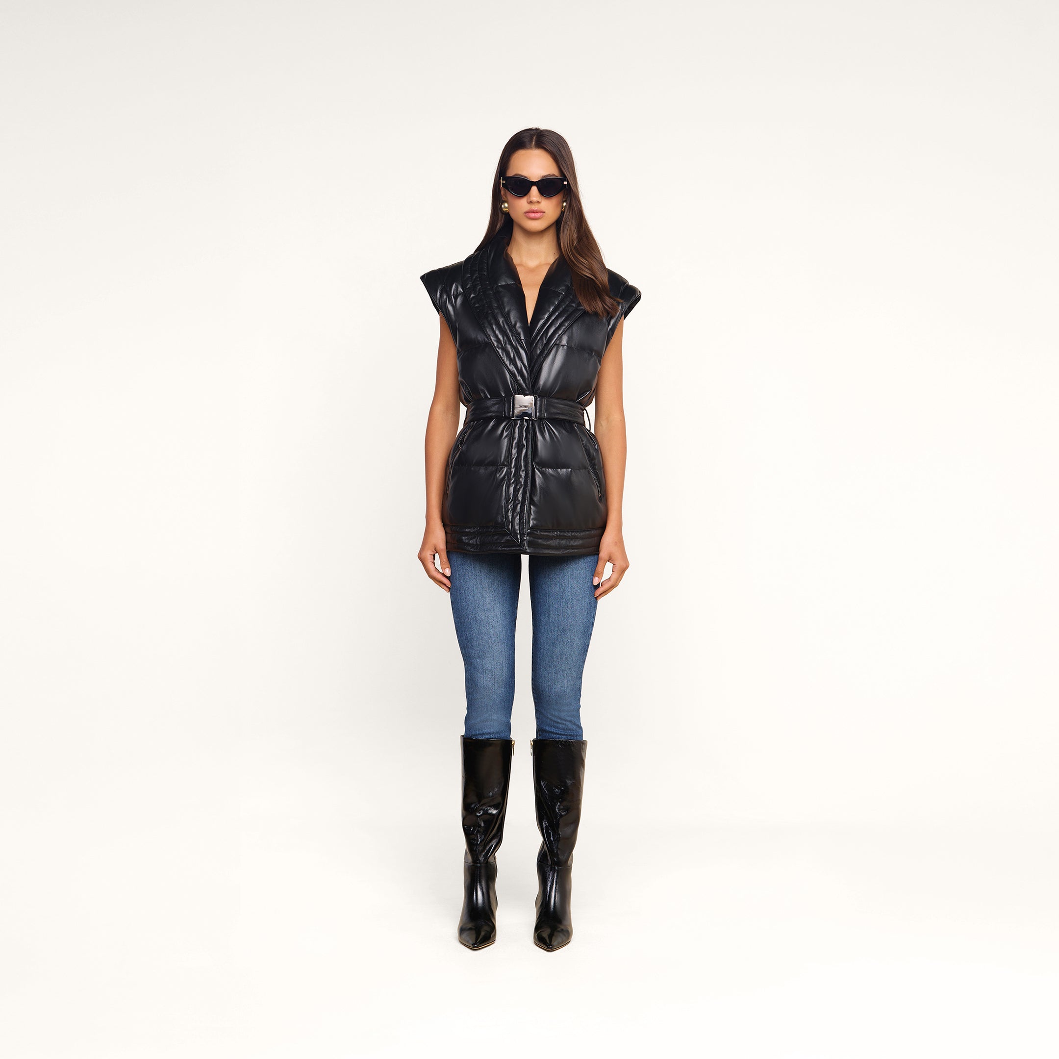 Wearing sunglasses, blue skinny jeans, and black knee-high boots, a woman stands facing forward against a plain white background in the Ignis Puffer Vest—a black vegan leather belted puffer with a shawl collar. This look is perfect for transitional outerwear.