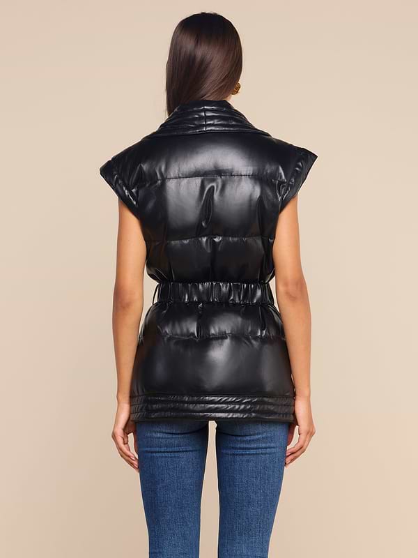 A woman stands facing away, wearing the Ignis Puffer Vest—a black, shiny vegan leather piece with a cinched waist—paired with blue jeans against a plain light background. This sleeveless vest is a versatile transitional outerwear option perfect for layering.