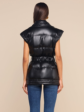 A woman with long brown hair, seen from behind, wears the Ignis Puffer Vest—a black, short-sleeve, belted vegan leather vest—paired with blue jeans for an effortlessly chic transitional outerwear look against a beige background.