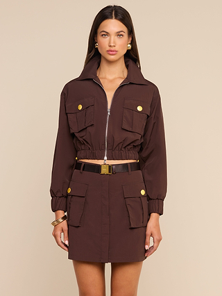 A woman wearing the Kade Windbreaker, a brown cropped utility-style jacket with large pockets and gold buttons, paired with a matching skirt, stands against a beige background.