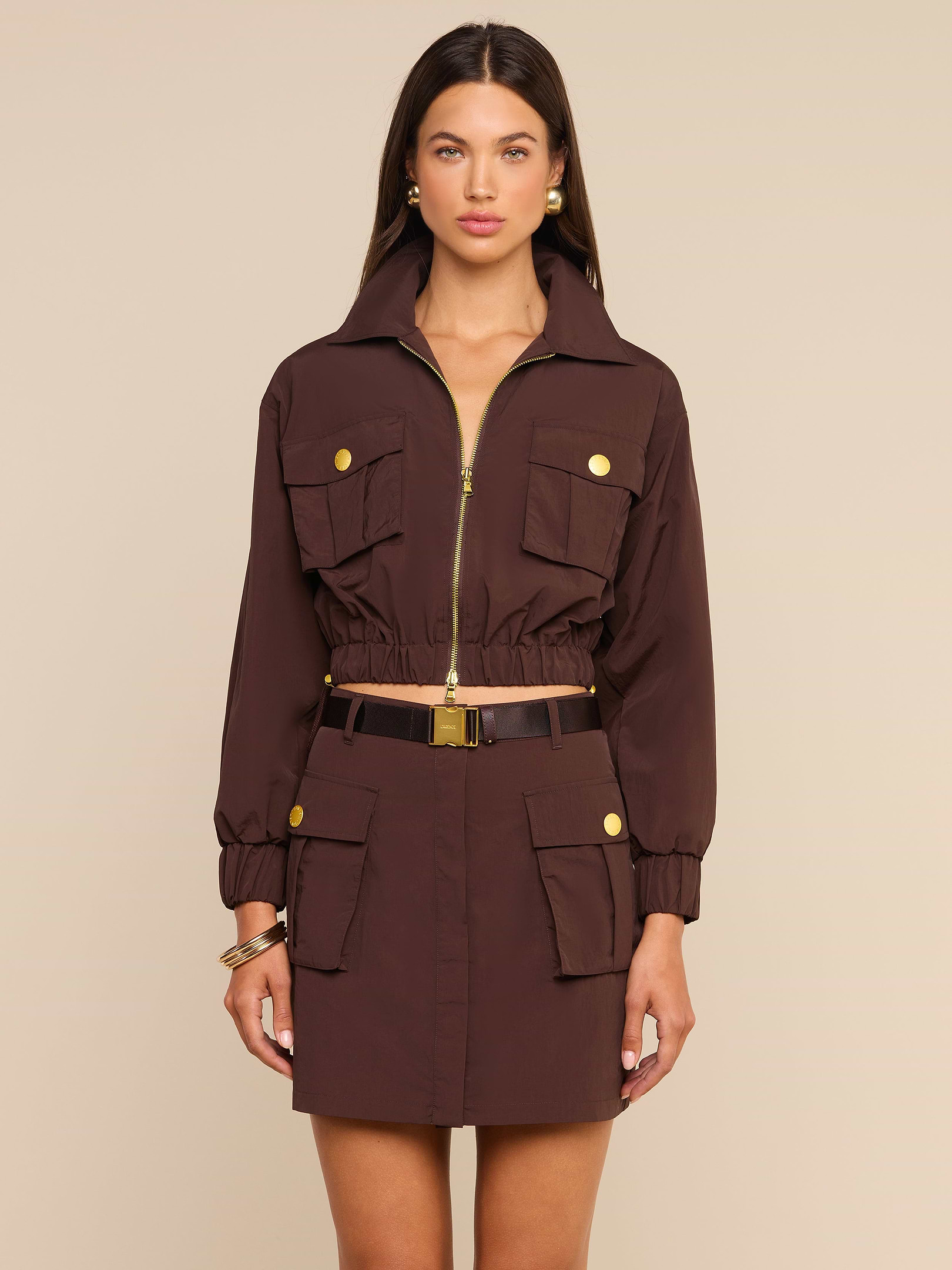 A woman stands facing forward wearing the Kade Windbreaker styled over a brown belted utility dress with gold buttons and multiple pockets, accessorized with gold jewelry against a plain light background.