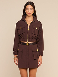 A woman stands facing forward wearing the Kade Windbreaker styled over a brown belted utility dress with gold buttons and multiple pockets, accessorized with gold jewelry against a plain light background.