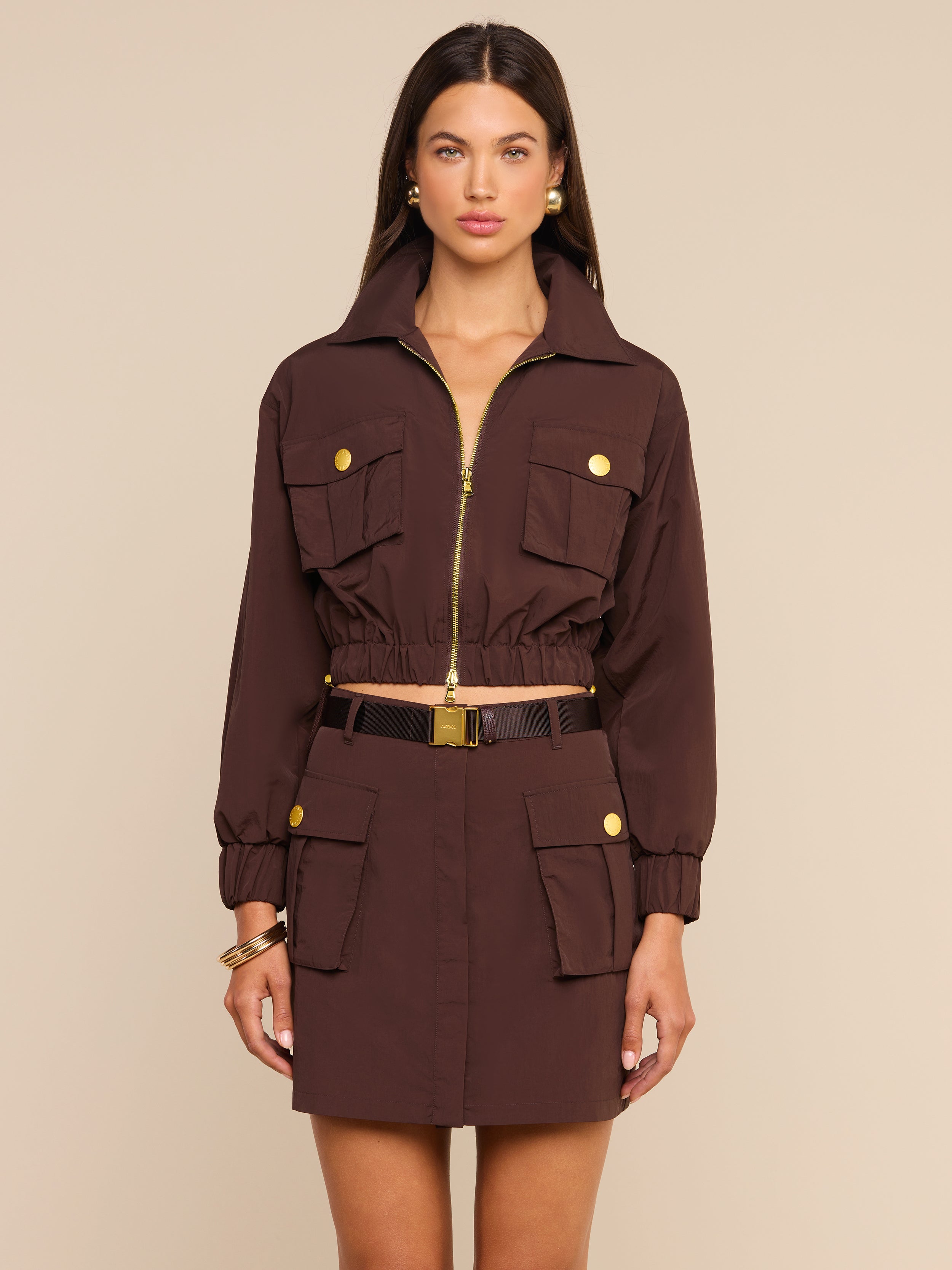 A woman stands facing forward wearing the Kade Windbreaker styled over a brown belted utility dress with gold buttons and multiple pockets, accessorized with gold jewelry against a plain light background.