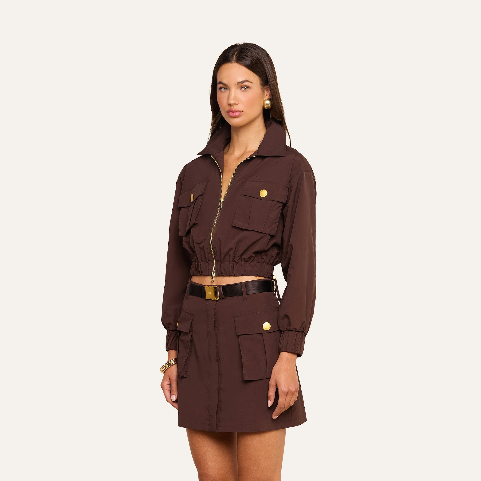 A woman wears the Kade Windbreaker, a brown cropped jacket with gold buttons, paired with a matching high-waisted mini skirt. She accessorizes her outfit with a black belt, gold earrings, and a bracelet while standing against a plain light background.