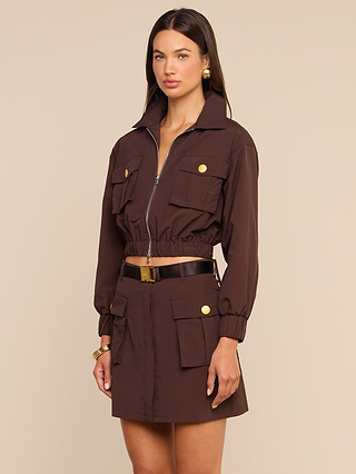 A woman stands against a beige background wearing the Kade Windbreaker—a brown transitional utility-style jacket with a cropped waist—paired with a matching skirt featuring gold buttons, gold earrings, and a black belt.