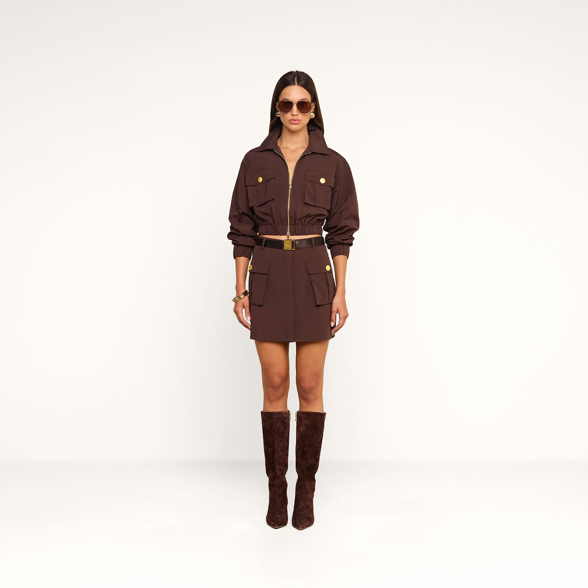 A woman wearing the Kade Windbreaker over a brown belted dress with large pockets, paired with matching brown knee-high boots, sunglasses, and gold jewelry, stands against a plain white background.