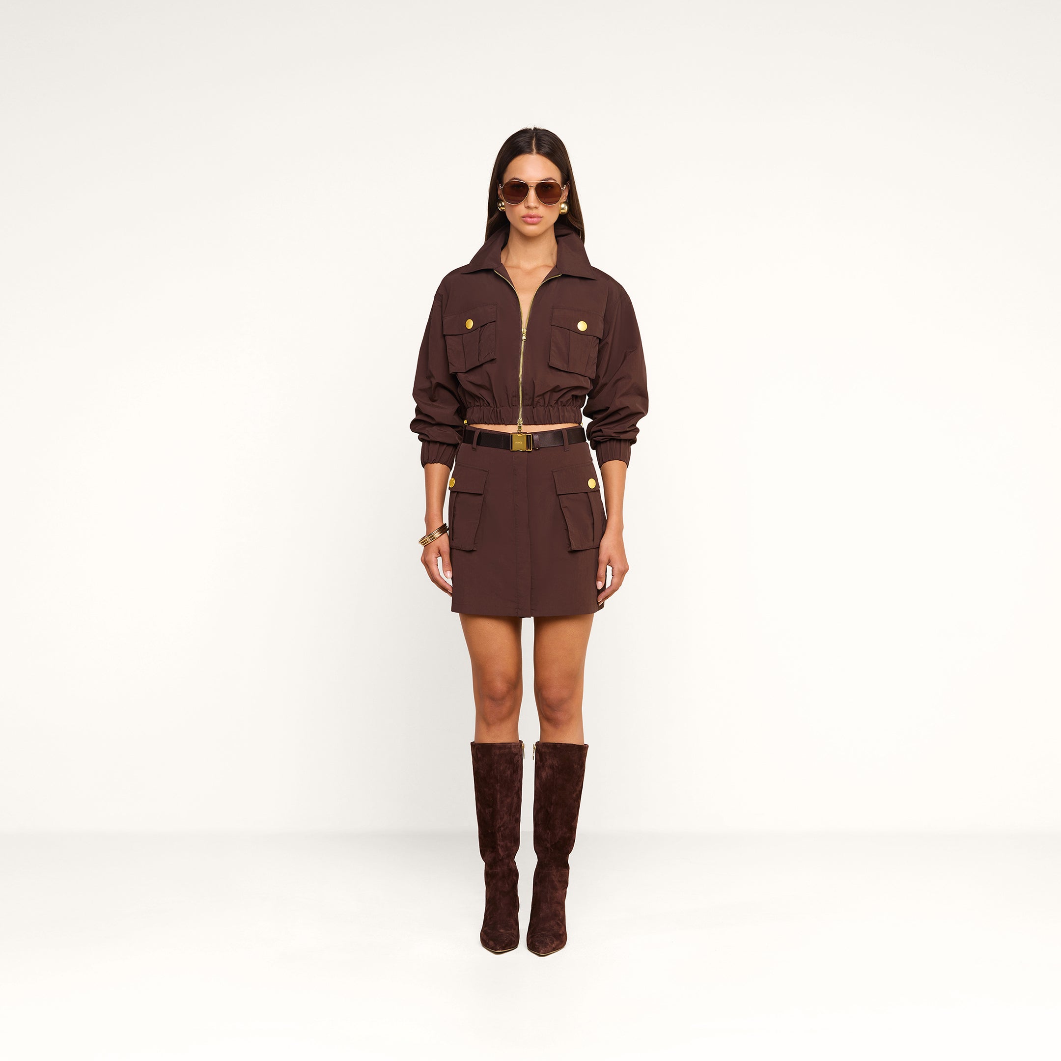 A woman wearing the Kade Windbreaker over a brown belted dress with large pockets, paired with matching brown knee-high boots, sunglasses, and gold jewelry, stands against a plain white background.