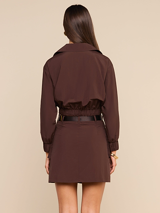 A woman with long brown hair stands facing away, wearing the Kade Windbreaker—a brown transitional jacket with a cropped waist—paired with a matching skirt against a beige background.