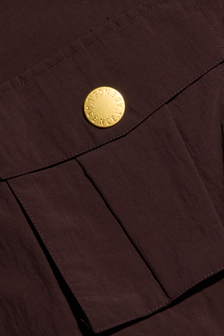 A close-up of a dark brown fabric pocket with a flap, secured by a round gold button embossed with text, highlights the refined detailing of the Kade Windbreaker.