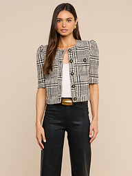 A woman stands against a plain background wearing the Allegra Cropped Tweed Jacket—a black-and-white plaid short-sleeve tweed piece with gold buttons—layered over a white top, paired with black high-waisted pants featuring a gold-buckled belt.