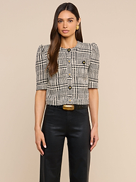 A woman with long brown hair is wearing the Allegra Cropped Tweed Jacket, which features a plaid pattern and short sleeves, paired with black pants, gold earrings, and a gold-buckled belt, standing against a plain light background.