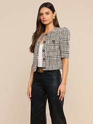 A woman stands against a plain background, wearing the Allegra Cropped Tweed Jacket with gold buttons over a white top, styled with black leather pants, a black belt, and gold earrings.