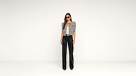 A woman wearing sunglasses, a checked short-sleeve top, and high-waisted black pants stands against a plain white background.